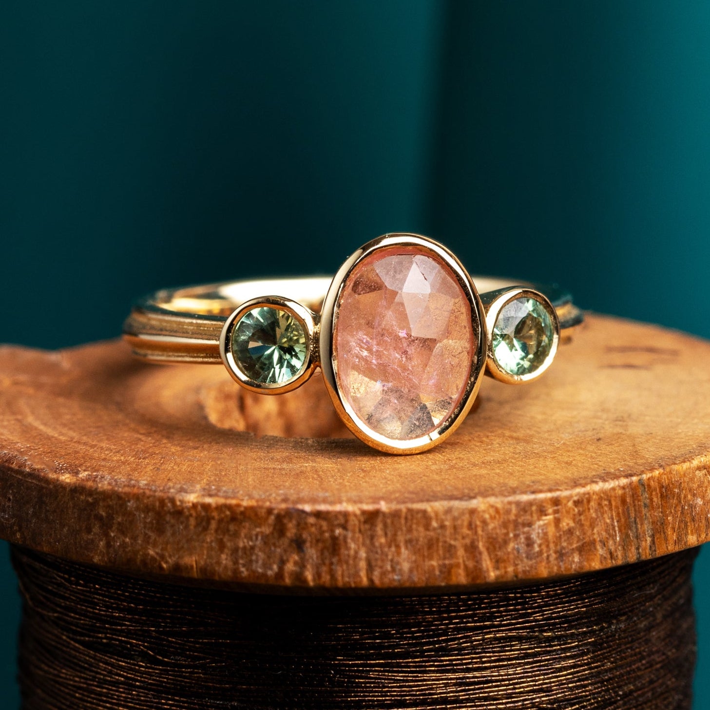 Roman style gold ring with a pink tourmaline and green sapphires on a wooden spool against a teal background