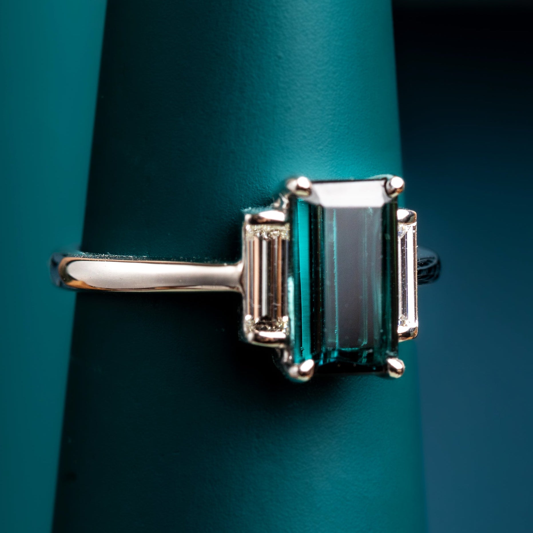 Platinum ring with a Indicolite Tourmaline and baguette diamonds on a teal stand against a dark background
