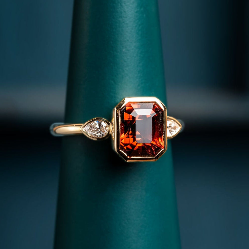 Gold ring with a large red orange tourmaline and pear shaped diamonds on a green stand.